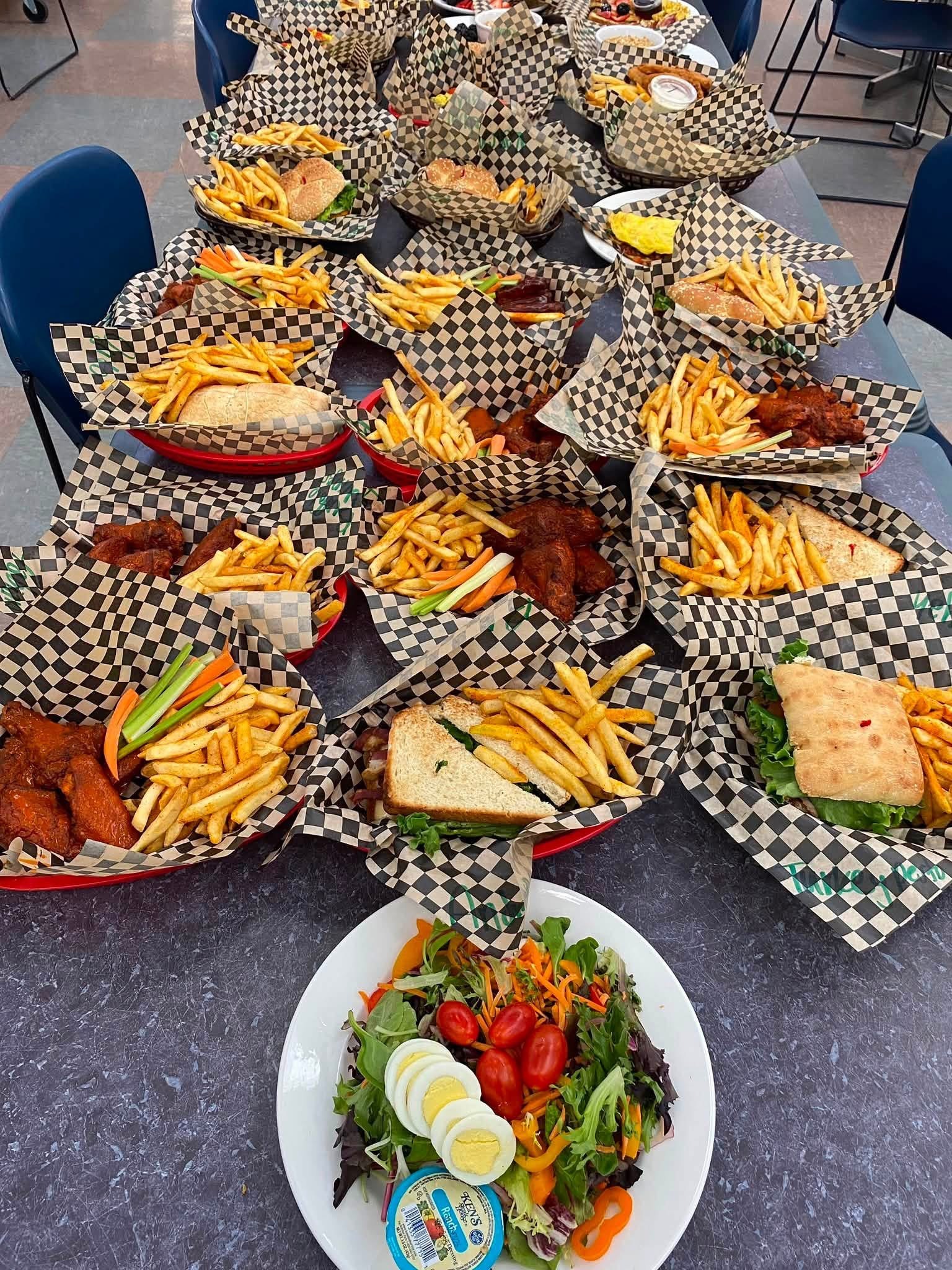 A wide table of catered sandwiches, wings, fries, and salad